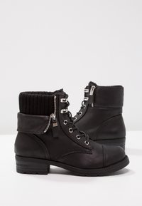 Paire de bottines en cuir noir avec lacets, fermetures éclair et poignets en tricot côtelé sur une surface blanche.