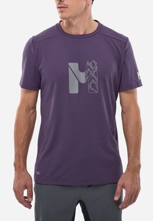 Homme portant une chemise à manches courtes violette avec un logo géométrique en forme de "M" sur la poitrine et une boucle d'oreille en forme de croix, debout devant un fond blanc.