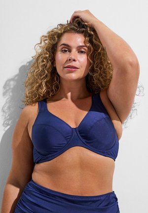 Kučaťavá žena, ktorá má na sebe tmavomodrú športovú podprsenku a zodpovedajúce spodné diely, pričom si jednu ruku opiera o hlavu na jednoduchom bielom pozadí.