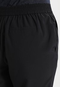 Černé kalhoty s elastickým pasem a hladkou texturou, se bočními kapsami a volným, pohodlným střihem. Tkanina vypadá lehce a prodyšně.