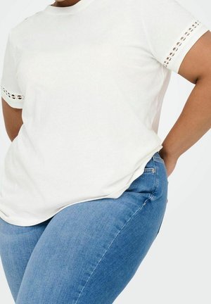 Personne portant une chemise blanche à manches courtes avec un détail œillet sur les manches et un jean en denim bleu, les mains reposant sur les hanches sur un fond uni.