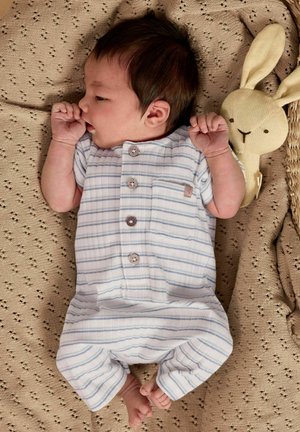 Naujagimis kūdikis dryžuotame baltame rūbelyje guli ant megztos smėlio spalvos antklodės, šalia galvos – smėlio spalvos pūkuotas zuikutis, žįsdamas nykštį.