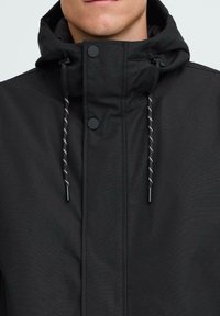 Črna jakna z visokim ovratnikom in nastavljivo kapuco, z gladko teksturo, pritisnimi gumbi in črno-belimi vrvicami.