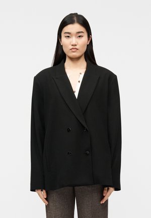 Ung kvinde med langt sort hår iført en oversize sort dobbeltradet blazer over en cremefarvet top og brune bukser, stående mod en ensfarvet baggrund.