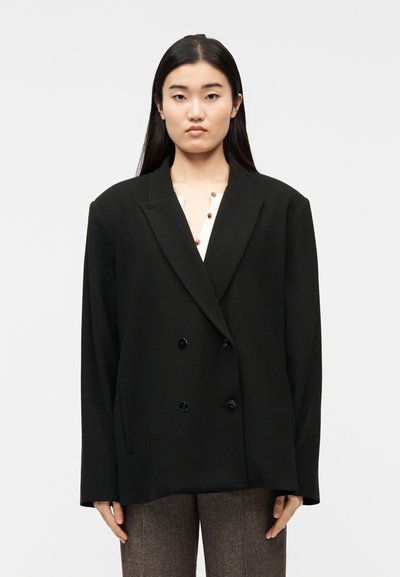 Jeune femme aux longs cheveux noirs portant un blazer noir surdimensionné à double boutonnage par-dessus un haut crème et un pantalon marron, debout devant un fond uni.