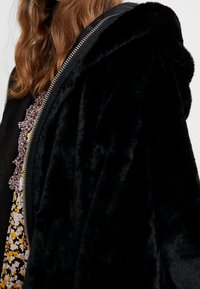 Czarna futrzana kurtka z kapturem, z suwakiem i miękką teksturą. Częściowo widoczny jest wzorzysty kwiatowy strój pod spodem.