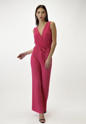 Fuchsia mouwloze jumpsuit met een diepe V-hals, geplooide textuur, strikceintuur en wijde benen. Zwarte sandaaltjes met bandjes maken de look compleet.