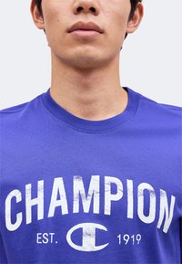 Νεαρό άτομο φοράει μωβ μπλουζάκι Champion με λευκό κείμενο που δείχνει το όνομα της μάρκας και "EST. 1919" από κάτω.
