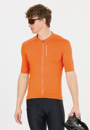 Jeune homme portant un maillot de cyclisme orange et un short noir, tenant un casque noir, regardant vers la gauche, portant des lunettes de sport réfléchissantes.