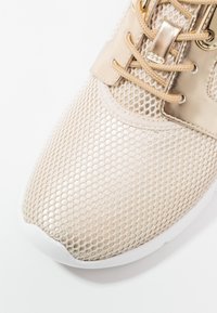 Jasnobeżowy but sportowy z siateczkową cholewką, gładkimi skórzanymi akcentami i białą gumową podeszwą. Złote metalowe elementy i sznurowadła.