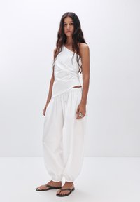 Witte mouwloze top met een asymmetrische halslijn en gedrapeerd detail, gecombineerd met witte, volumineuze, geraffineerde broeken en zwarte slippers.