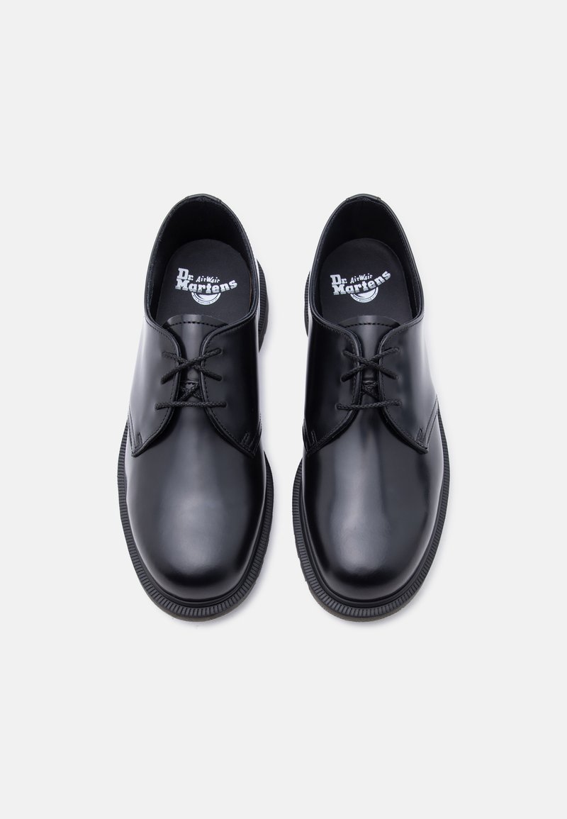 Paar schwarze, polierte Leder Dr. Martens Schuhe mit drei Ösen, abgerundeten Zehen und dicken Gummisohlen auf weißem Hintergrund.