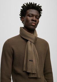 Écharpe en tricot marron avec des bords frangés, fabriquée en matériau doux, drapée de manière décontractée autour du cou sur un pull marron texturé.