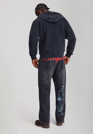Mann trägt schwarzen Hoodie, schwarze Jeans mit Schlangen- und Totenkopf-Design am rechten Bein und braune Schuhe, steht mit dem Rücken zu einem grauen Hintergrund.