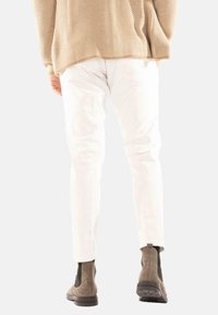 Persoon in witte broek, een beige gebreide trui en grijze suède Chelsea-boots, van achteren gezien tegen een effen achtergrond.