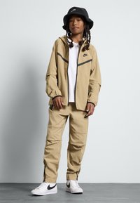 Conjunto de jaqueta e calças Nike bege com detalhes pretos, material leve, bolsos com fecho e um logótipo no peito e na perna. Inclui sapatilhas brancas.