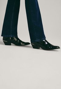 Stivali neri in pelle fino alla caviglia con punta affilata e tacco basso impilato, abbinati a jeans in denim blu scuro, che mostrano un aspetto elegante e rifinito.