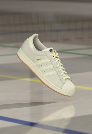 Basket Adidas blanche avec "MESSI" en lettres dorées sur le côté, semelle gomme et bout coqué, suspendue au-dessus d'un sol de terrain de sport.