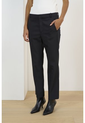 Osoba nosząca czarne, dopasowane spodnie do kostek, z ręką w kieszeni oraz czarne, szpiczaste botki na obcasie, stojąca na jasnej drewnianej podłodze.