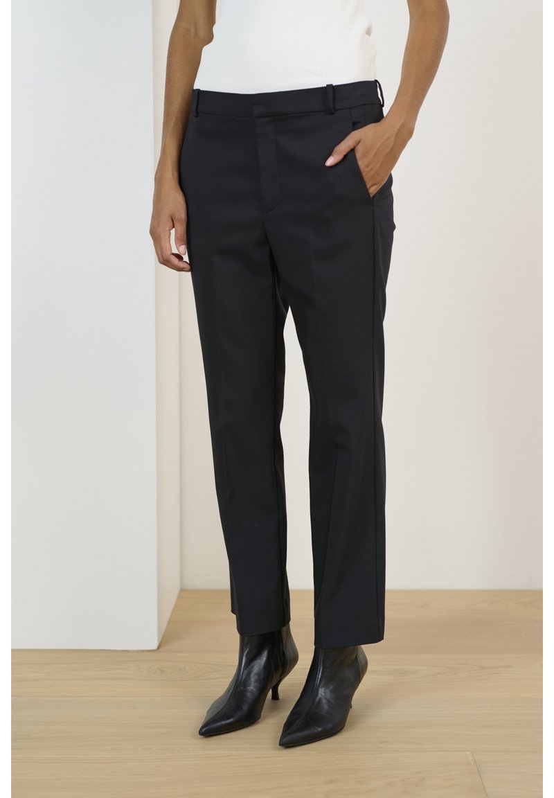 Personne portant un pantalon noir sur mesure, longueur cheville, main dans la poche, et des bottines noires à talons pointues, debout sur un sol en bois clair.