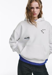Witte hoodie met een voorkantvak, voorzien van zwarte stergrafieken op de borst en de tekst "Urban Myth" aan de zijkant.