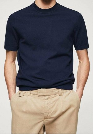 Mand iført en navyblå kortærmet sweater stoppet ned i beige plisserede bukser med hænderne i lommerne, står mod en neutral baggrund.