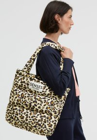 Padded tote bag in a beige and brown leopard print, featuring a thick strap and a branded label, providing a textured, spacious design.