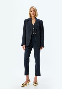 Blazer a righe navy con rever a punta, abbinato a un gilet coordinato e pantaloni su misura. Tacchi dorati con fiocchi completano l'outfit.