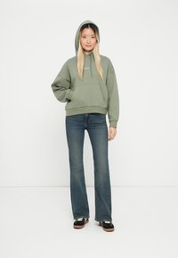 Groene capuchontrui met een voorzak, gecombineerd met donkerblauwe flared jeans en zwarte sneakers. Model staat tegen een effen achtergrond.