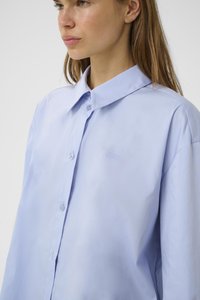 Camicia azzurra chiaro con colletto ampio, realizzata in tessuto morbido. Presenta un logo ricamato discreto sul petto e una vestibilità comoda.