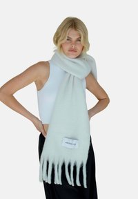 Écharpe en laine gris clair avec des bords frangés et une étiquette de logo, enroulée autour du cou, portée sur un débardeur blanc.