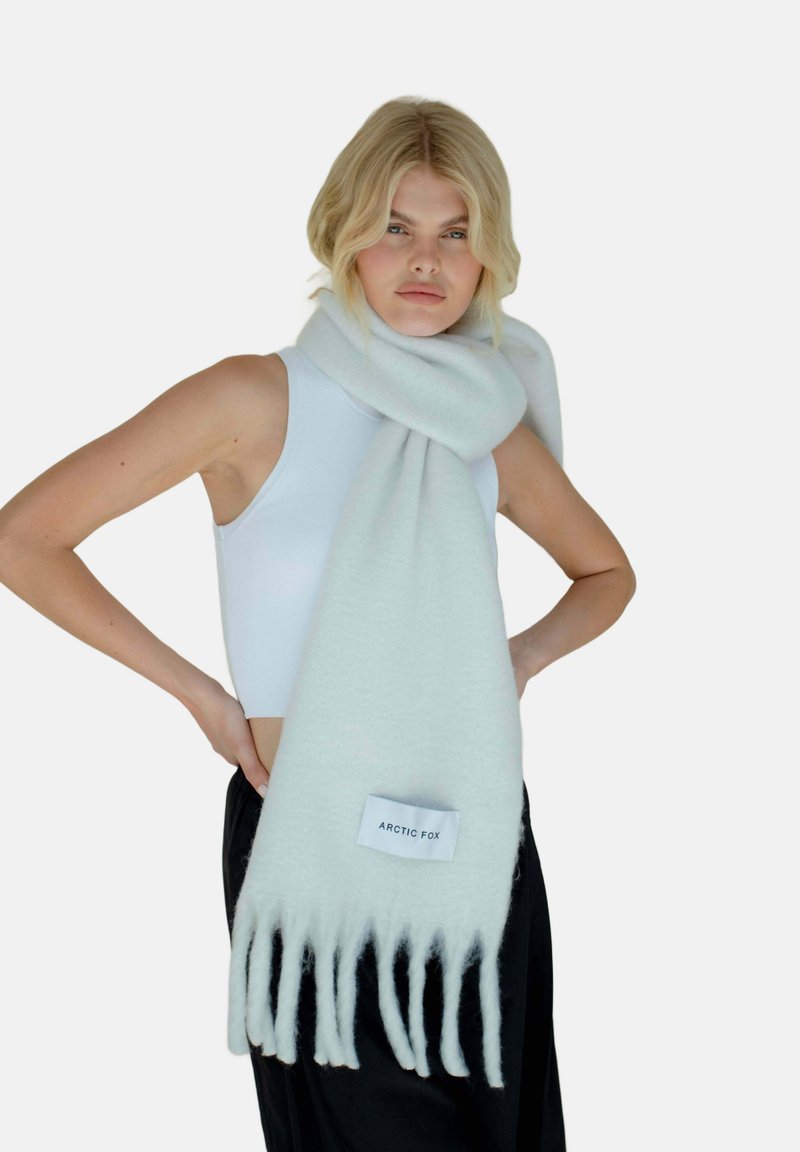 Écharpe en laine gris clair avec des bords frangés et une étiquette de logo, enroulée autour du cou, portée sur un débardeur blanc.