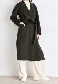 Abrigo de lana verde oliva con cintura ceñida, grandes bolsillos, solapas con muescas y un diseño largo y estructurado, combinado con pantalones blancos de canalé.