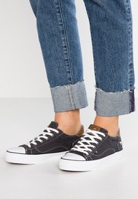 Černé plátěné tenisky s bílými gumovými podrážkami, bílými tkaničkami, hnědými koženými akcenty a prošívanými detaily. Ohrnuté džíny ukazují lemy.