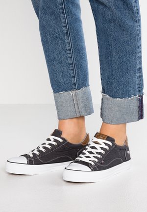 Zwarte canvas sneakers met witte rubberen zolen, voorzien van witte veters, bruine leren accenten en gestikte details. Gekrulde spijkerbroek met zichtbare zomen.