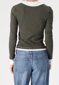 Maglietta a maniche lunghe verde oliva con texture a coste, indossata sopra una camicia bianca; abbinata a jeans in denim azzurro chiaro con tasche posteriori.
