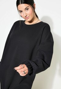 Mustan ylimitoitettu collegepaita, jossa on ribbidettu kaulus ja kerätty hihansuu. Pehmeä kangas, rento istuvuus, jossa on sileä pinta ja minimalistinen muotoilu.