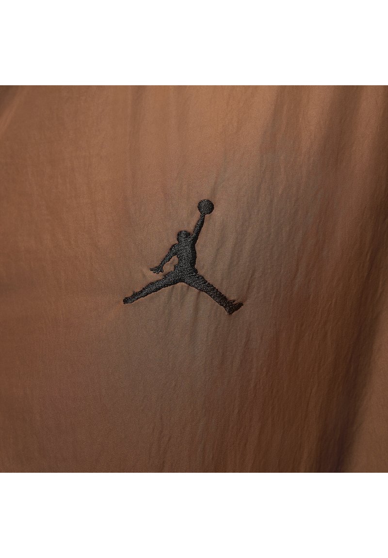 Schwarze Silhouette eines Basketballspielers, der springt und mit einem ausgestreckten Arm einen Ball hält, auf braunem Stoff gestickt.