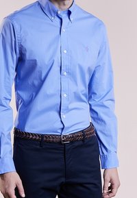 Ljusblå skjorta med krage, prydd med en liten lila logotyp på bröstet; matchad med ett mörkt vävt bälte och mörka byxor.