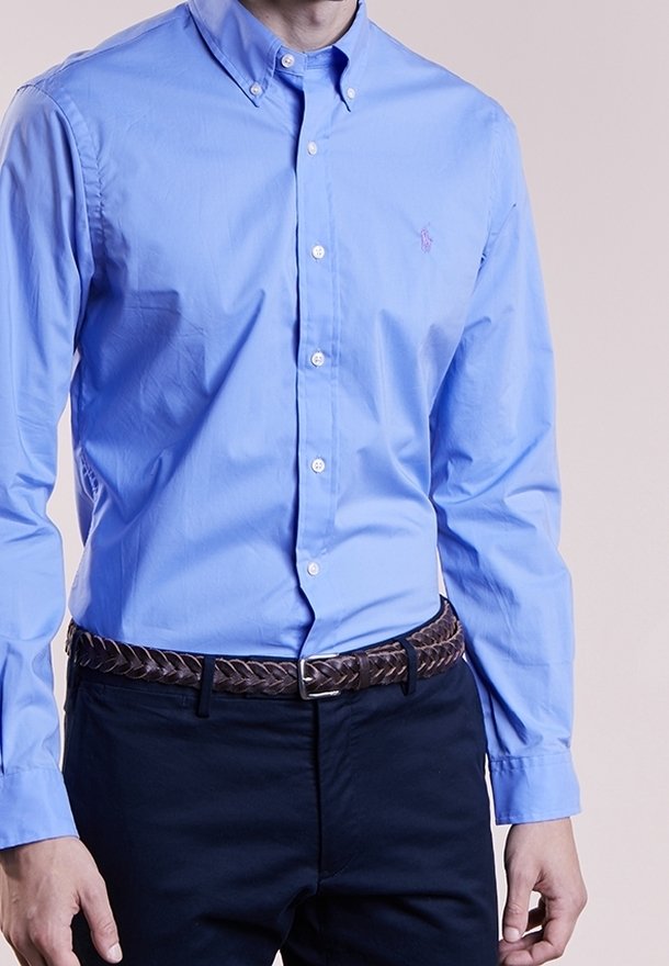 Ljusblå skjorta med krage, prydd med en liten lila logotyp på bröstet; matchad med ett mörkt vävt bälte och mörka byxor.