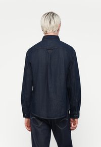 Denimskjorte i mørkeblå med lange ermer, spiss krage, knappemanchetter og synlig søm. Har en buet hem og tilbake yoke-detalj.