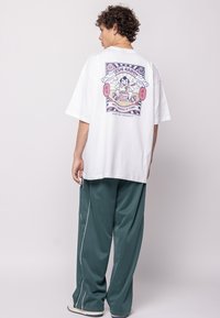White oversized t-shirt with a colorful graphic print featuring a noodle theme on the back. Paired with loose green pants with white piping.