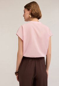 Blusa rosa a maniche corte con taglio dritto e scollatura rotonda, abbinata a pantaloni marroni con vita elastica e bracciali in legno.