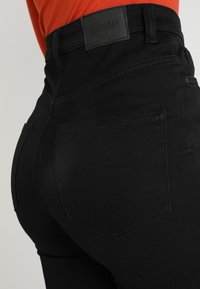 Jeans negros de tiro alto con un diseño ajustado, que cuentan con un bolsillo trasero y una etiqueta negra de "WEEKDAY" en la cinturilla.
