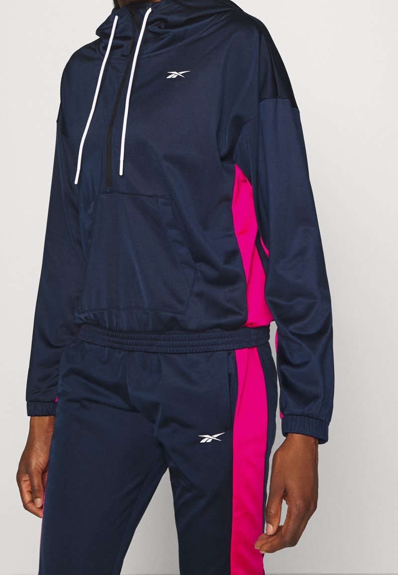 Personne portant un survêtement Reebok bleu marine et rose vif avec une veste à capuche et un pantalon, avec des cordons blancs et logos.