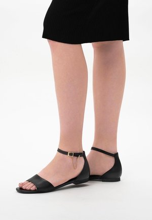 Schwarze Ledersandalen mit offenem Zehendesign. Verfügen über einen Knöchelriemen mit einem goldenen Kettenakzent und flacher Sohle. Glatte Textur, minimalistisches Design.