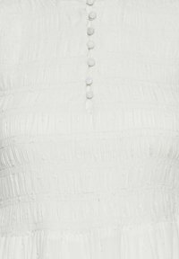 Biała sukienka z teksturowanej tkaniny, z pionowymi paskami i małymi, okrągłymi guzikami pokrytymi tkaniną, biegnącymi wzdłuż przodu.