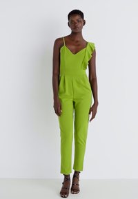 Žalias šokių kostiumas su prigludusia krūtine, V formos iškirpte, vieno peties volanu ir plonais, pritaikytais kojų kirpimais; lengvas audinys ir elegantiškas dizainas.