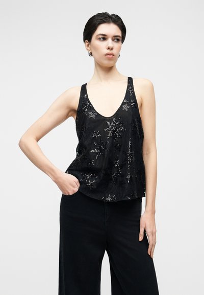 Črno majico brez rokavov z globokim V-izrezom, okrašeno s cvetličnimi vzorci iz bleščic. Mehka tkanina, brez rokavov. Kombinirana s črnimi širokimi hlačami.
