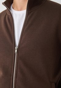 Hnedá zipsová bunda z hladkého materiálu, s vysokým golierom a strieborným zipsom, oblečená na bielom tričku pod ňou.